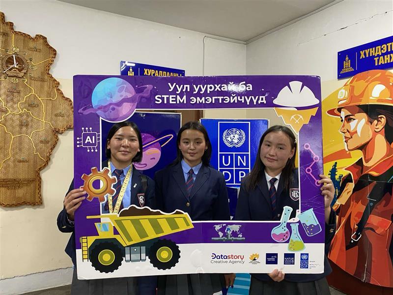 Four people pose behind a large purple STEM photo frame with UNICEF logos in a classroom.