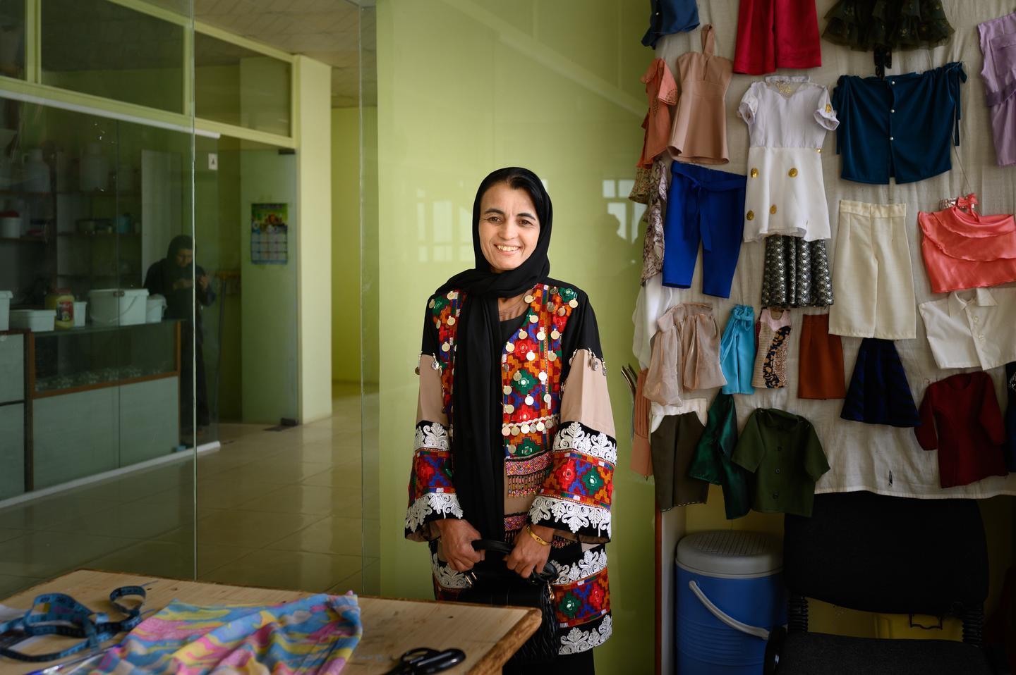 Smiling woman in a hijab wearing a colorful patchwork vest stands behind a fabric shop counter.