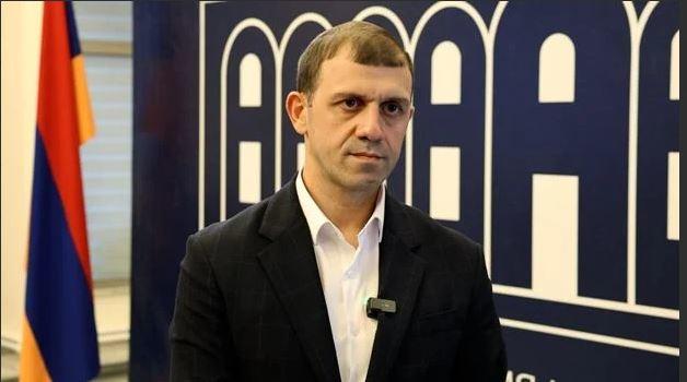 Man in a dark suit with lapel mic at a press event, blue backdrop with white letters and a flag nearby.