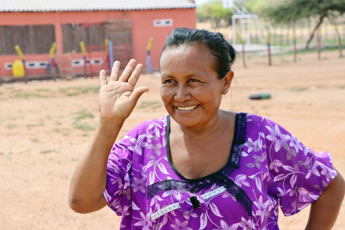 Person in a purple patterned shirt raises a hand in a dusty outdoor area.
