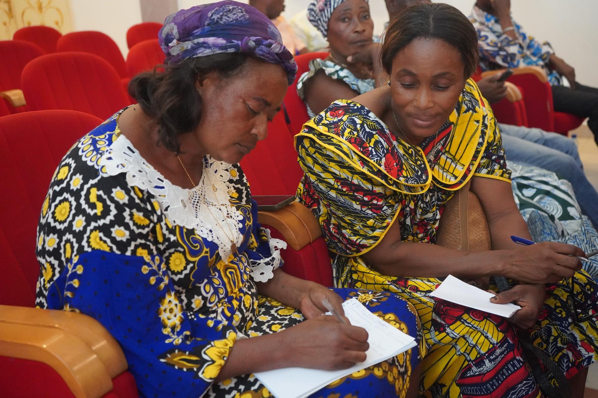 Two women in vibrant patterned dresses sit at red theater seats, taking notes.