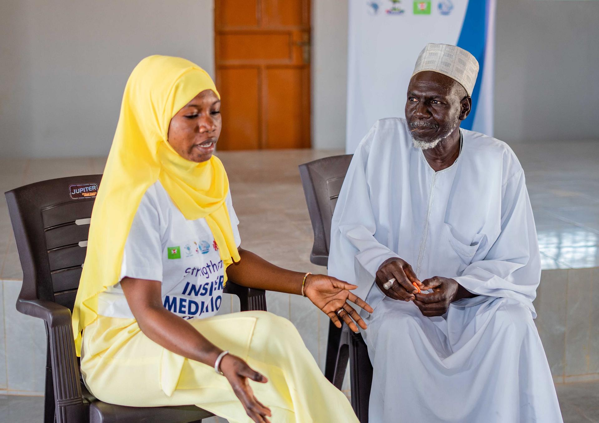 Smiling woman in a yellow hijab sits beside a man in white traditional robe indoors.
