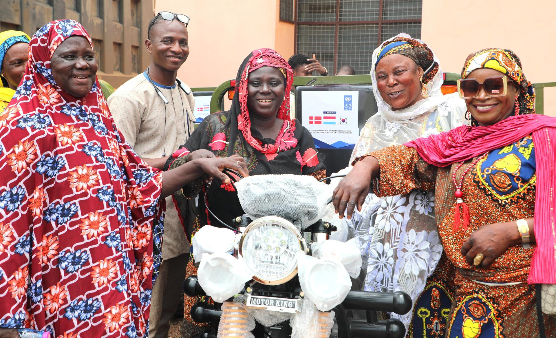 Group of people in colorful traditional clothing pose with white motorcycle beside peach building.