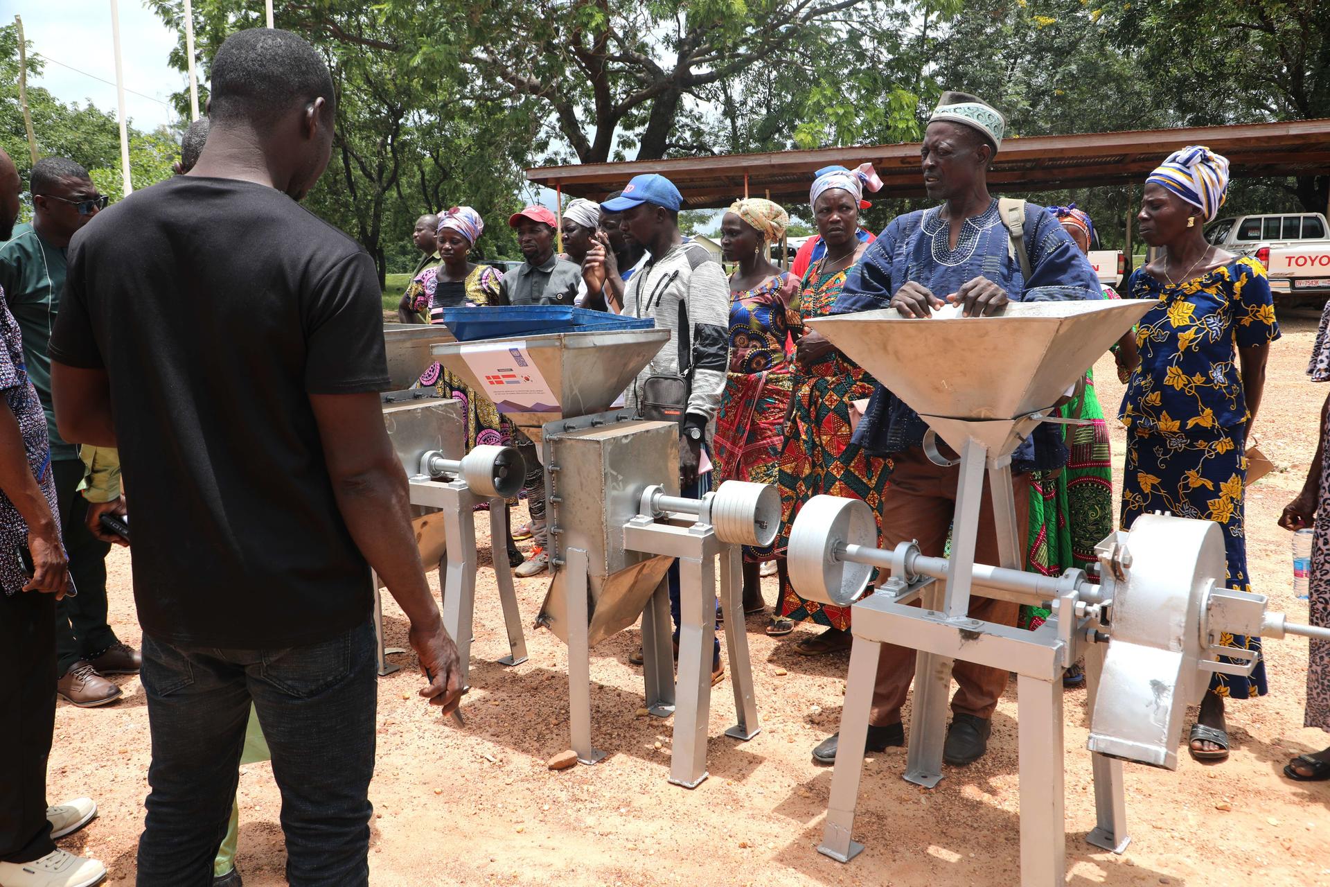 Group of people gathered around several hand-cranked grain mills outdoors.