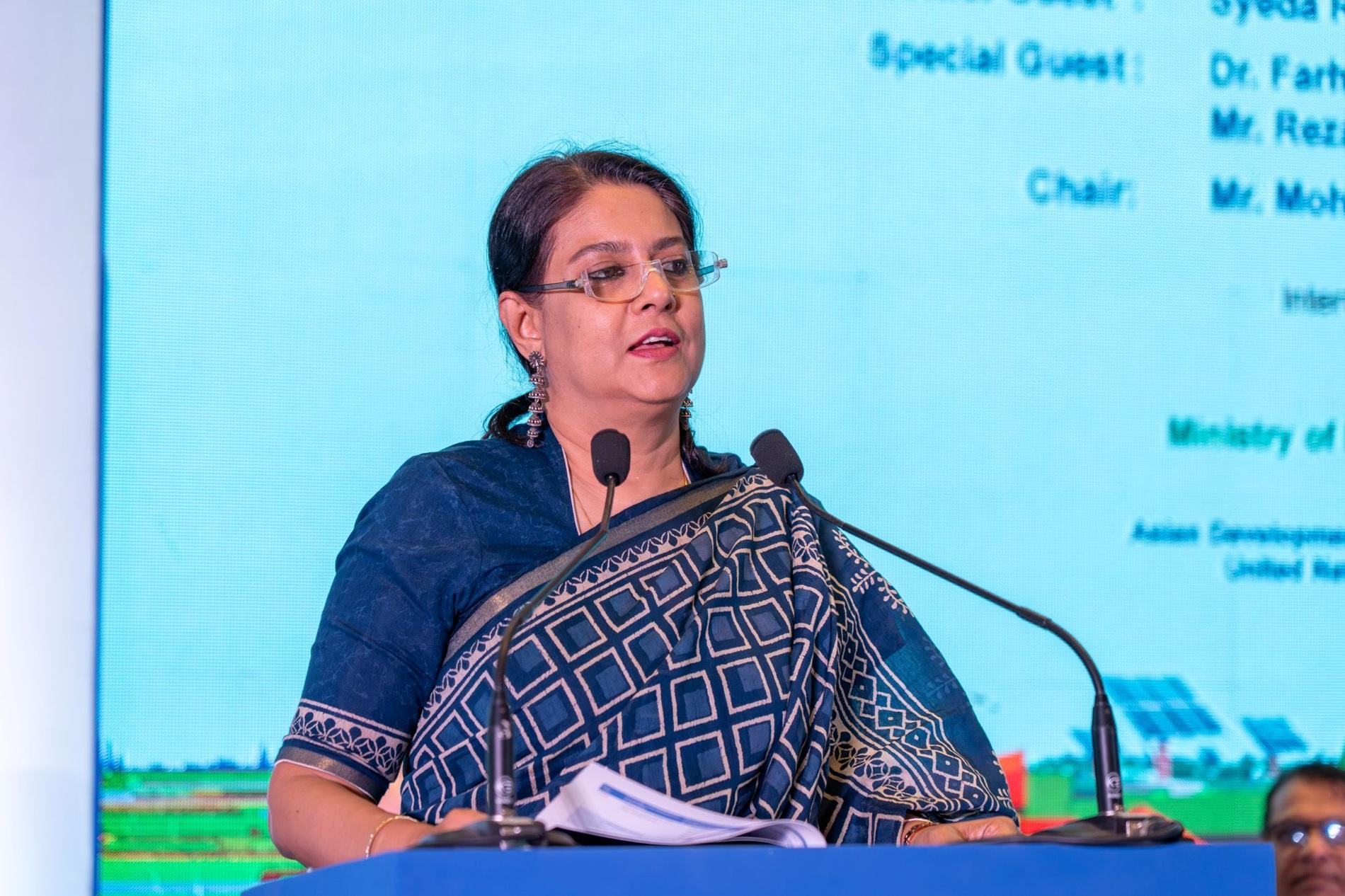 Speaker in a blue patterned outfit at a podium with a microphone, blue screen behind.