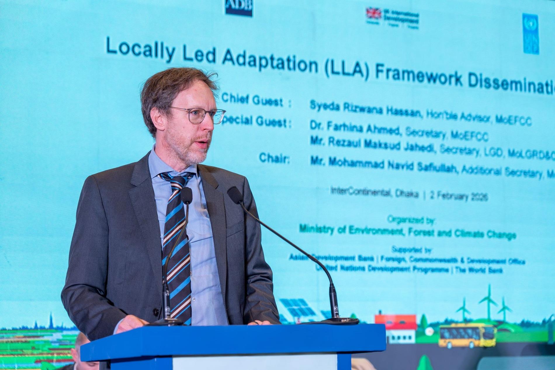 Speaker at podium on stage, blue screen displays LLA Framework Dissemination.