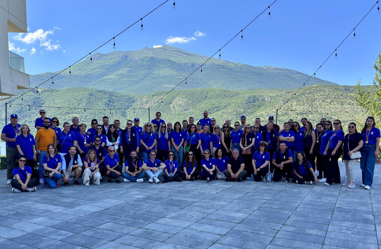 Large group in blue shirts posing for a photo on a patio with string lights and mountains behind.