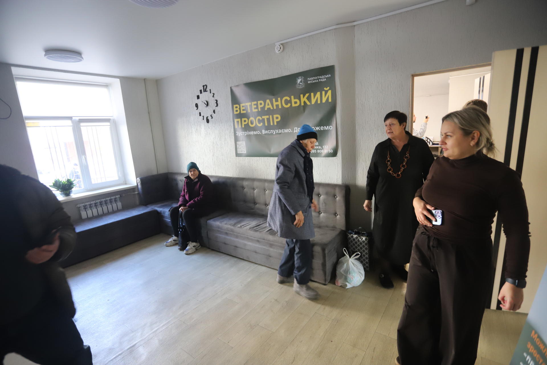 Indoor room with several adults standing around; window, a couch, and a banner on the wall.