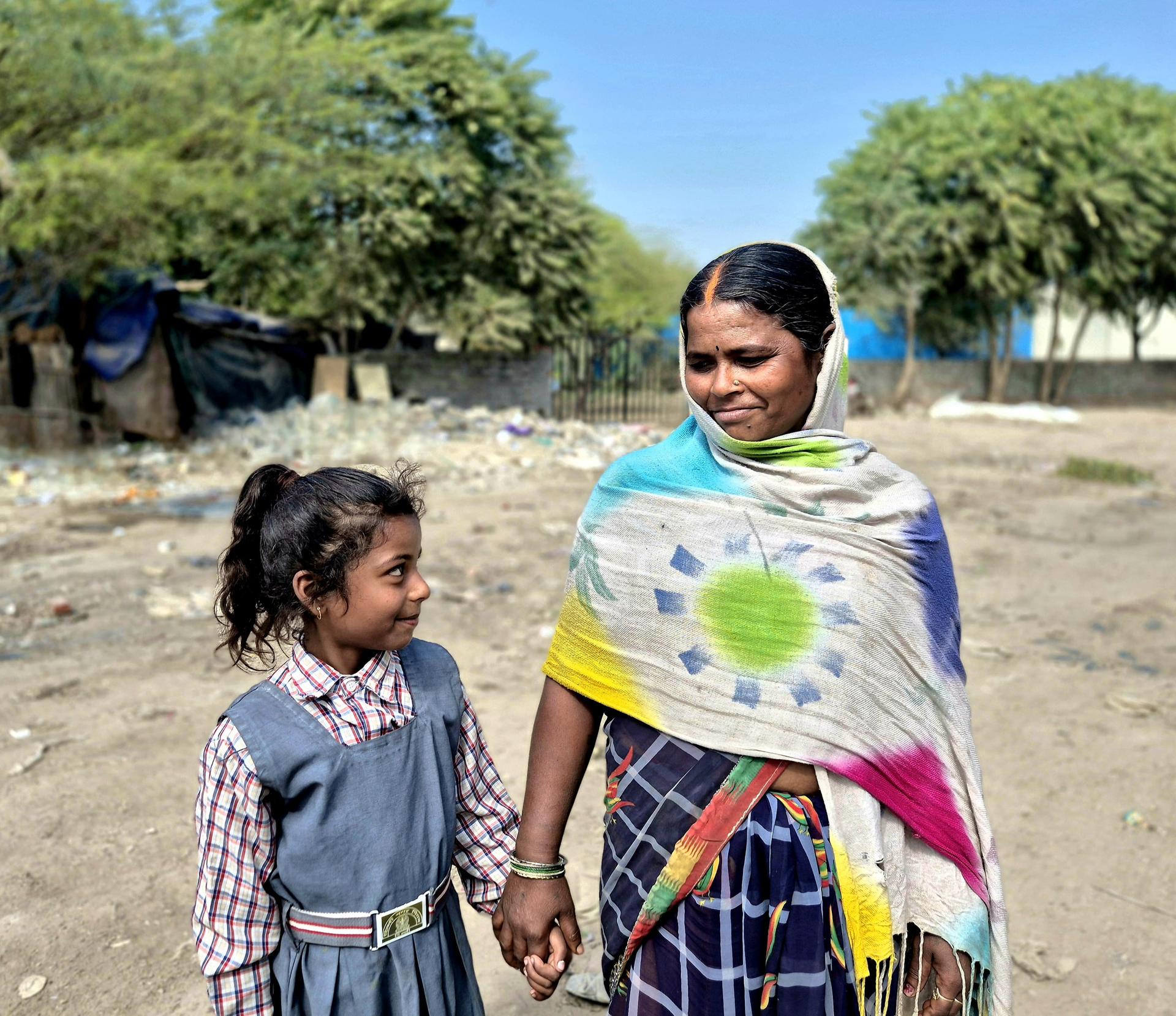 Two people holding hands outdoors on dusty ground; child in plaid dress, adult in colorful shawl.