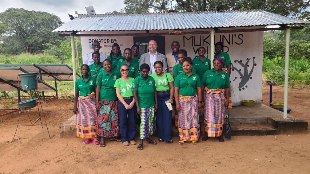 Group of people in green shirts posing under a canopy outdoors on dusty ground.