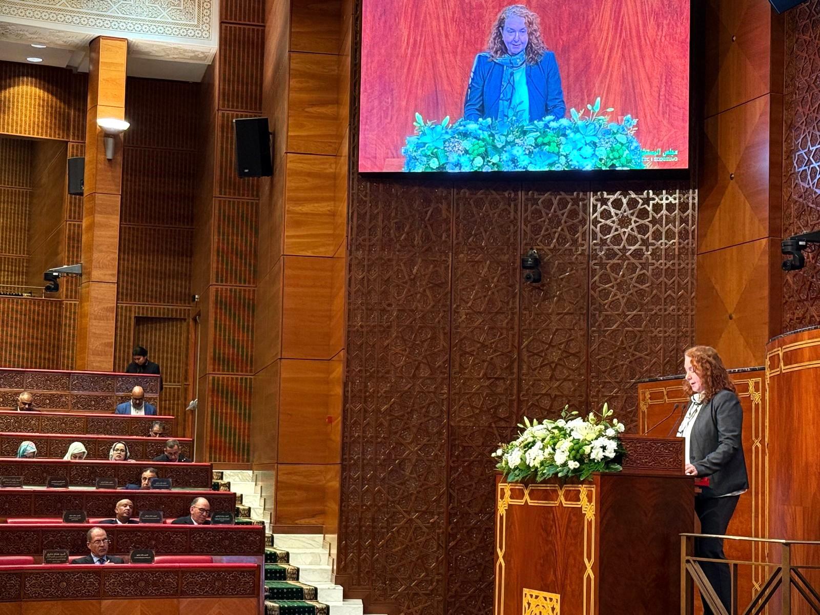 Lecture hall stage with a speaker at a flowered podium; audience seated, screen shows person in blue.