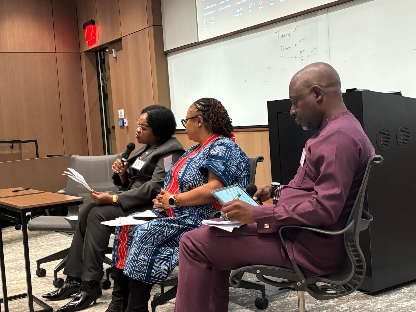 Three panelists seated on stage in a lecture hall, reading papers.