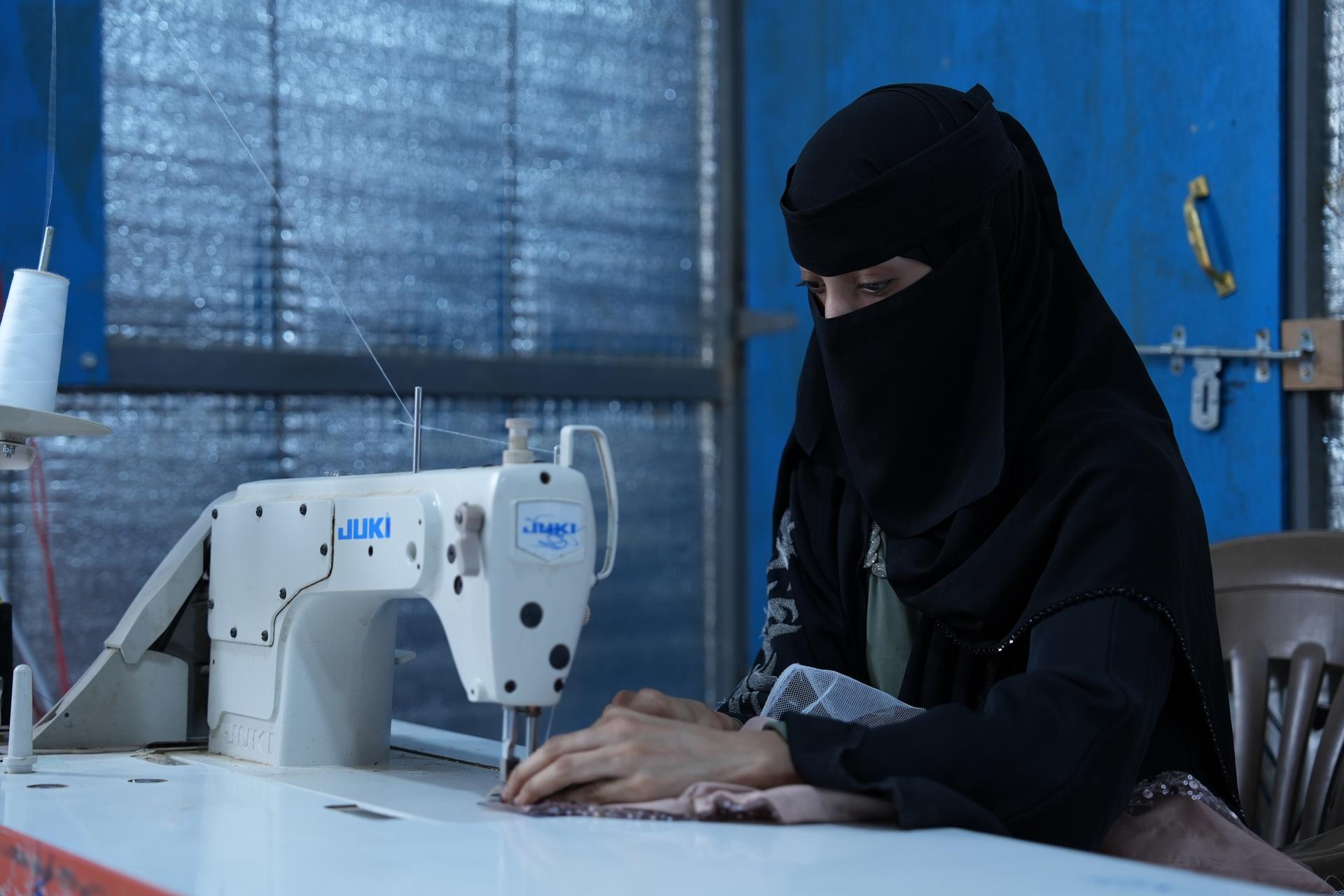Photograph: a woman in a black hijab sewing at a white sewing machine in a blue workshop.