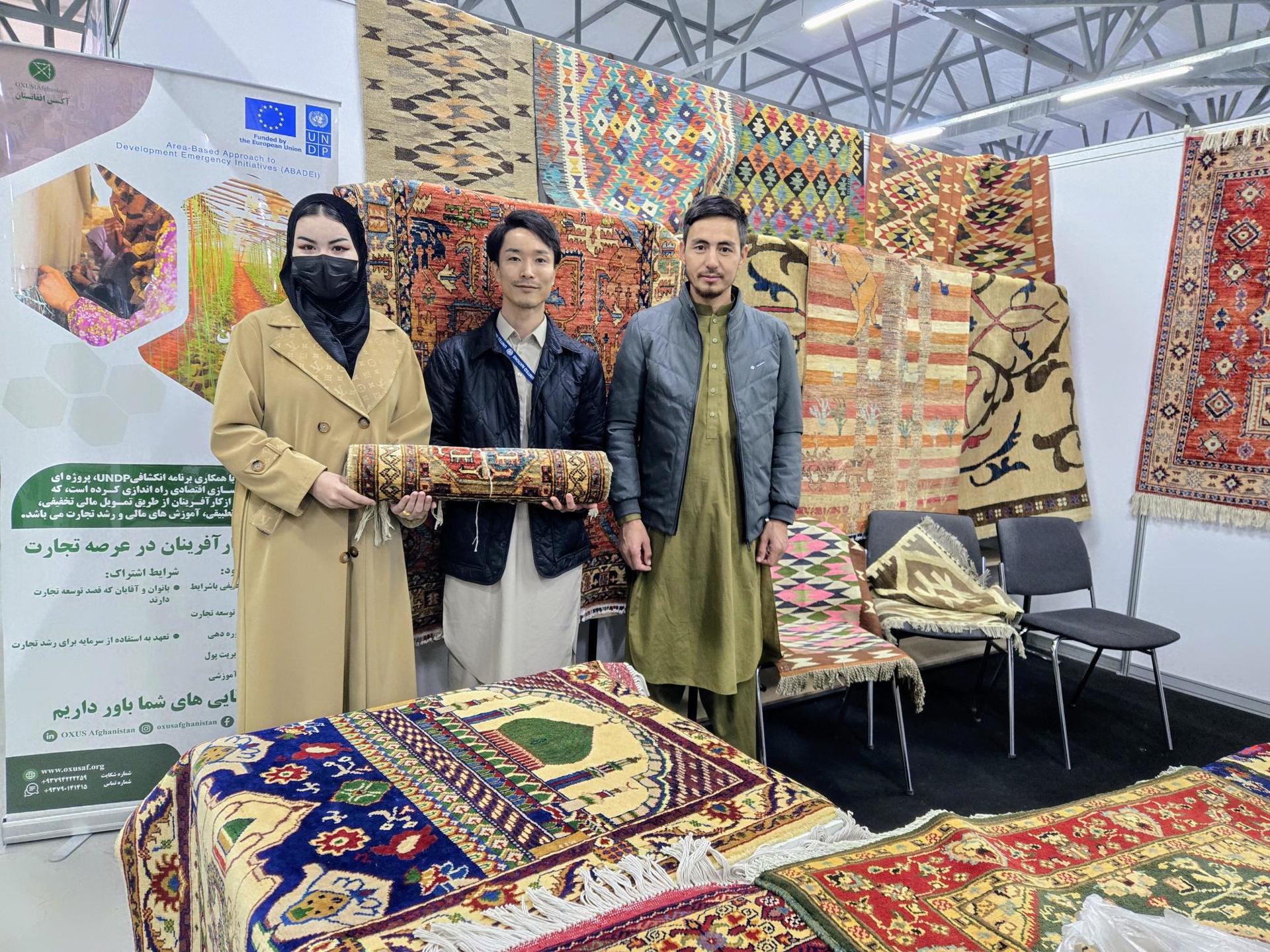 Four people in masks stand in a rug booth at a textile fair, colorful carpets on display.