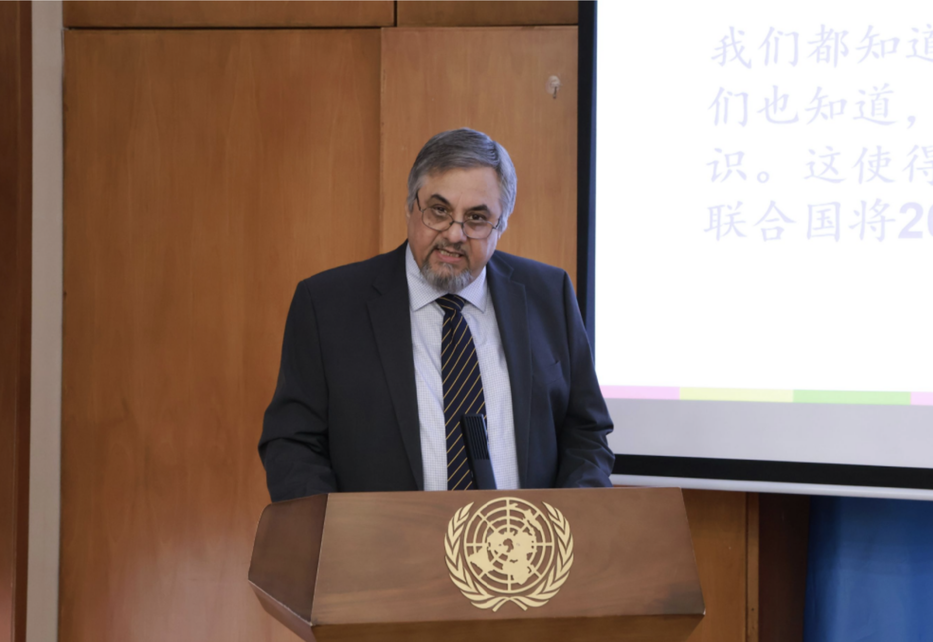 Speaker at podium with seal, presenting beside a projection screen showing Chinese text.