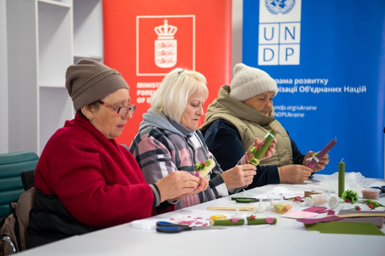 Three people at a table with colorful paper crafts; red crest and UNDP banners in back.