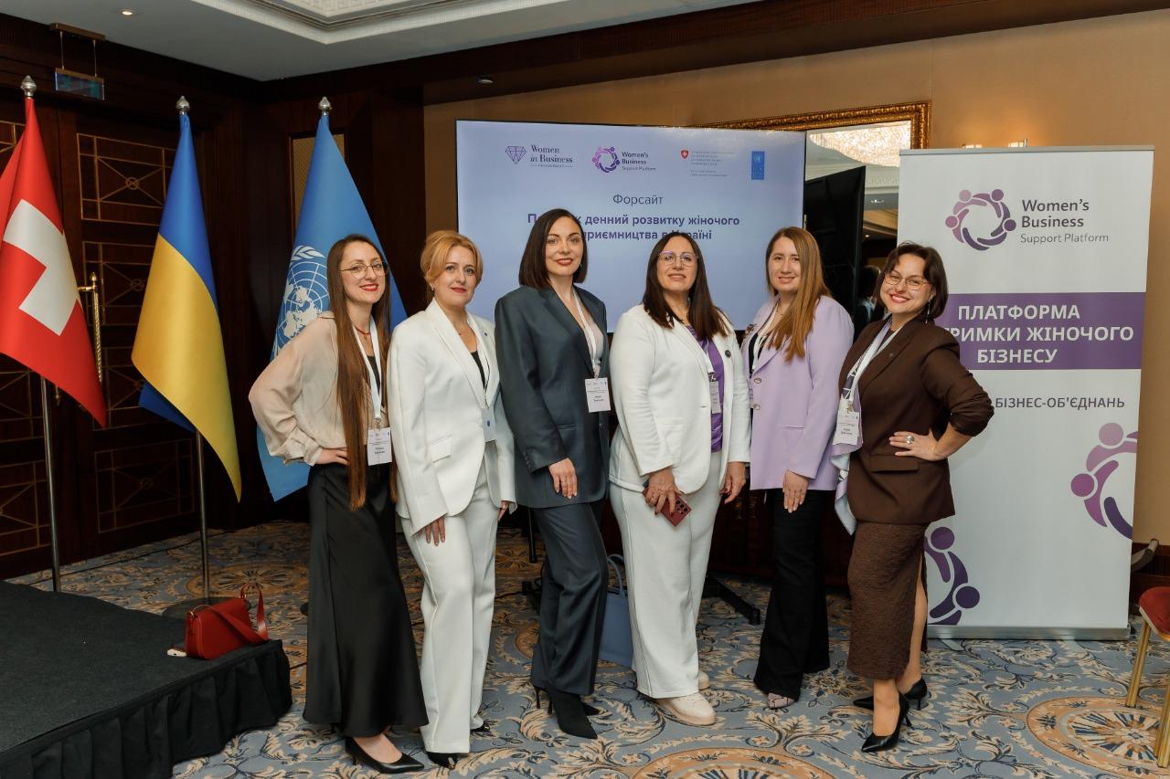 Group of women pose on a conference stage with blue banners and flags in the background.