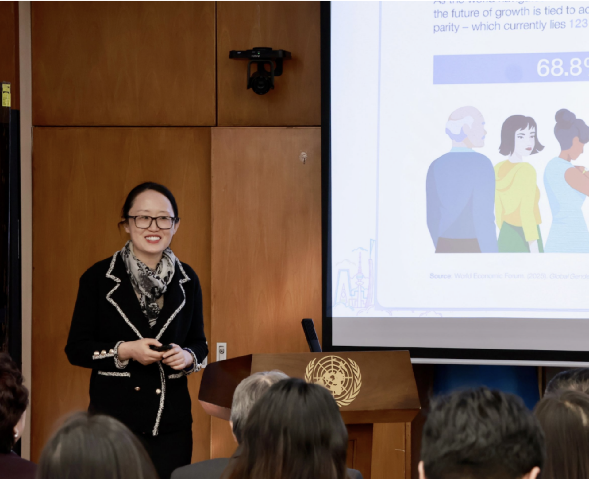 Photo: female presenter in black speaks to an audience in a classroom, with a projected slide.
