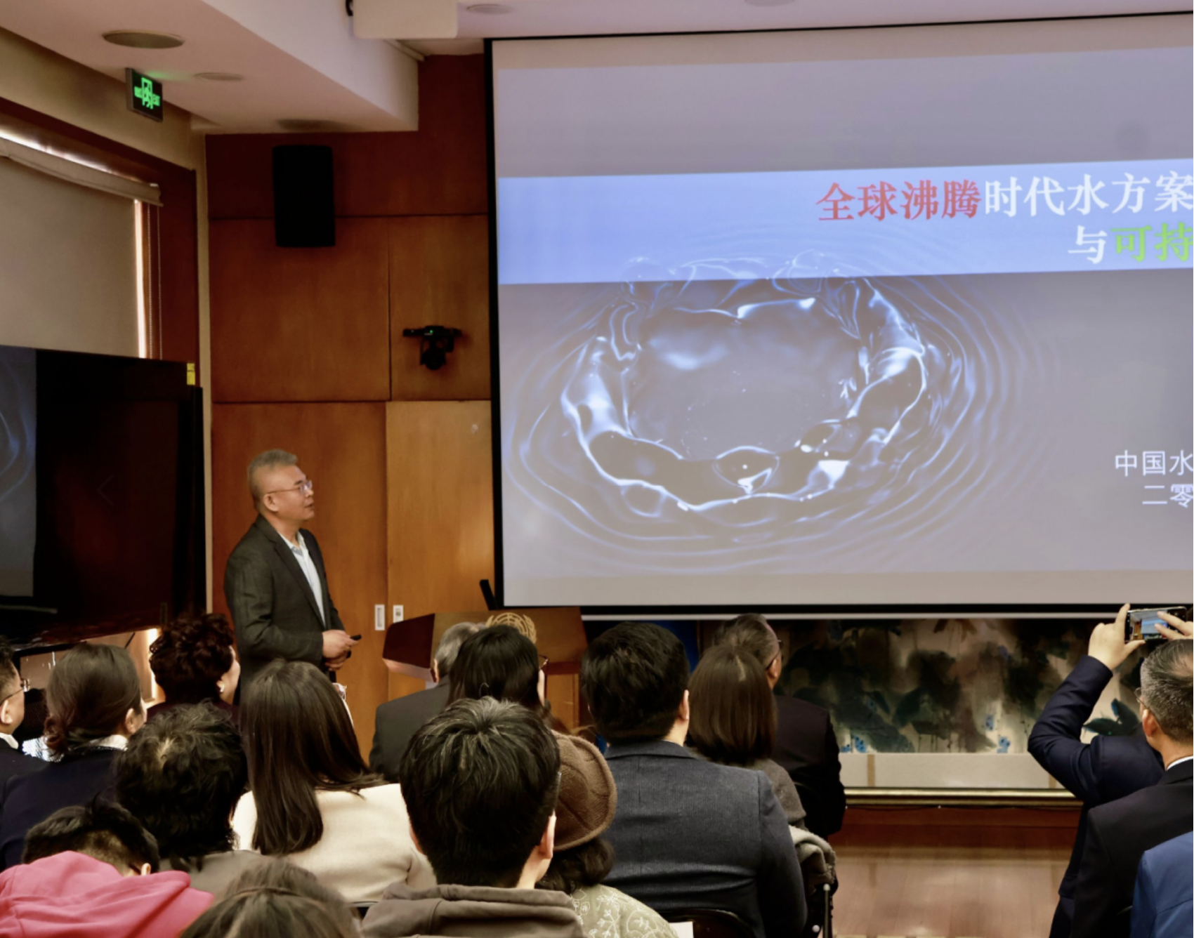 Photograph of a speaker presenting to an audience in a conference room; water-splash slide.