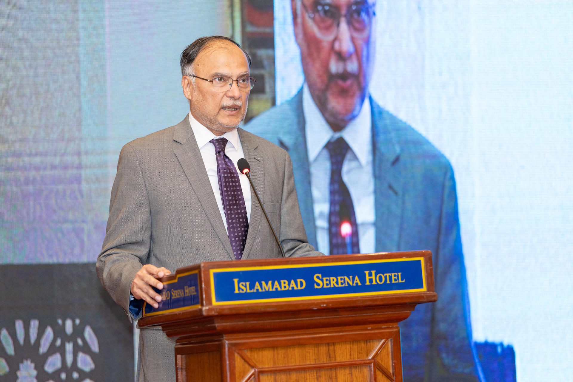 Speaker in gray suit at podium with Islamabad Serena Hotel sign; large screen behind.