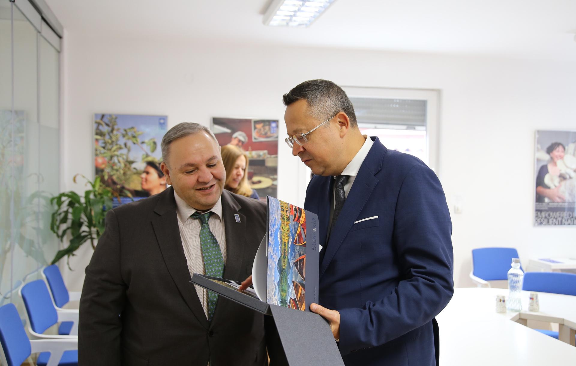Two suited men review a colorful brochure in a bright office conference room.