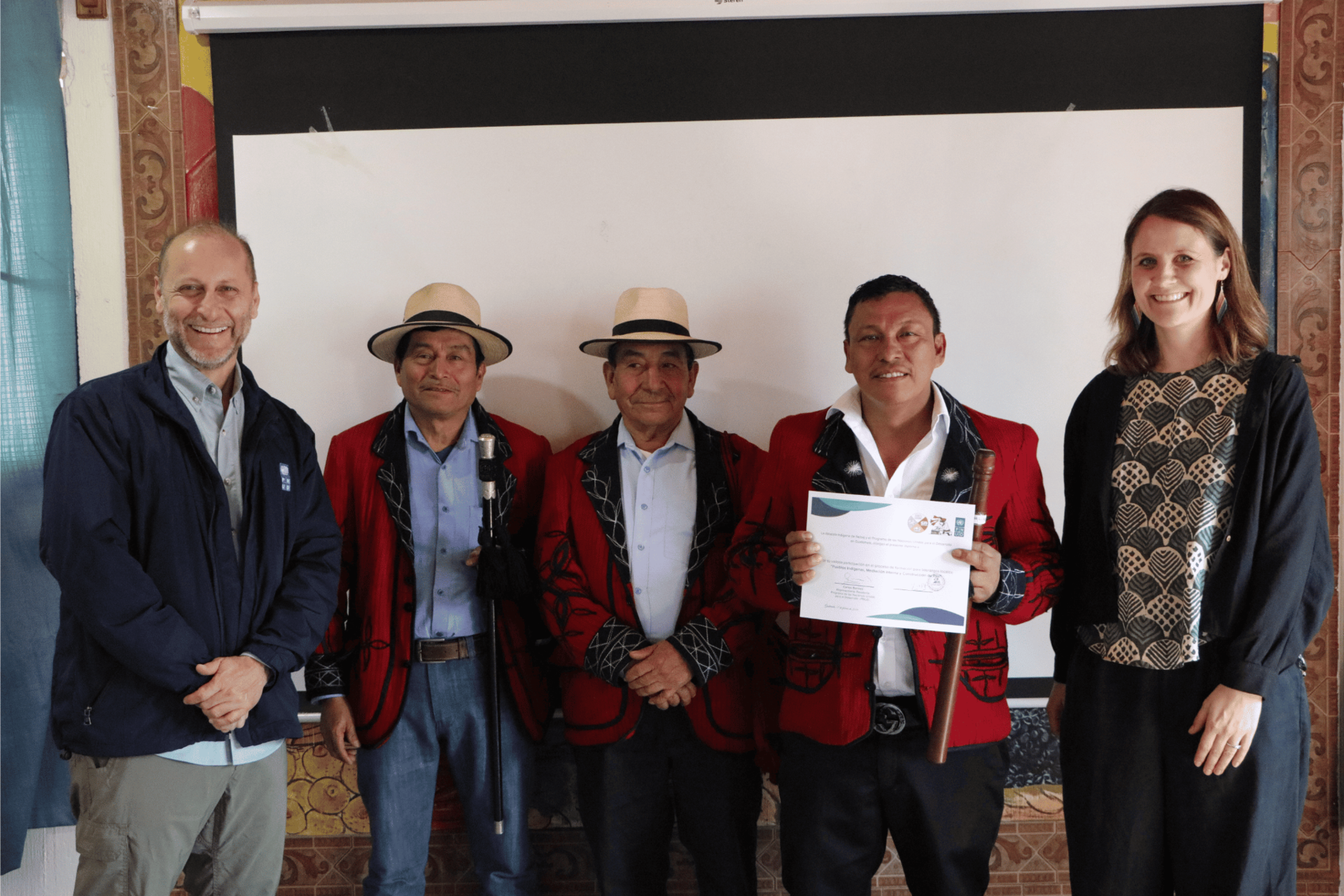 Six people pose indoors before a projection screen; three men wear red vests, one holds certificate.