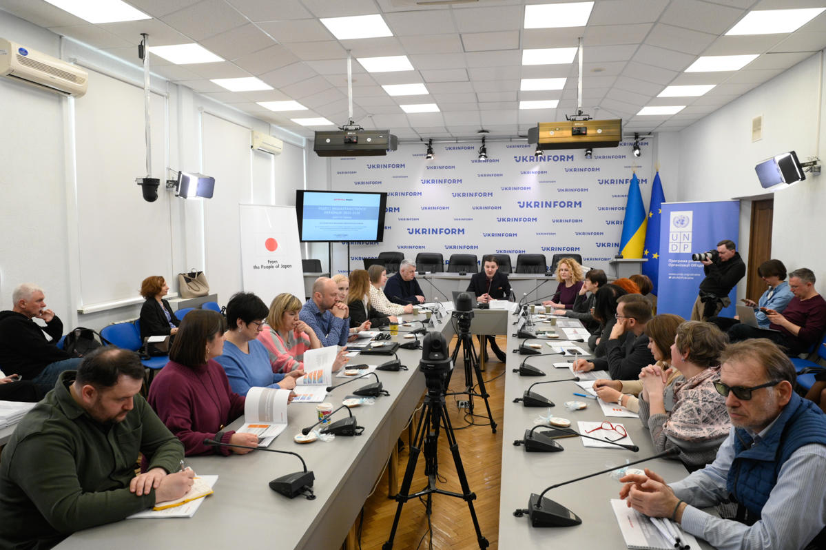 A large conference room with a long table of attendees, microphones, a screen, and the Ukraine flag.