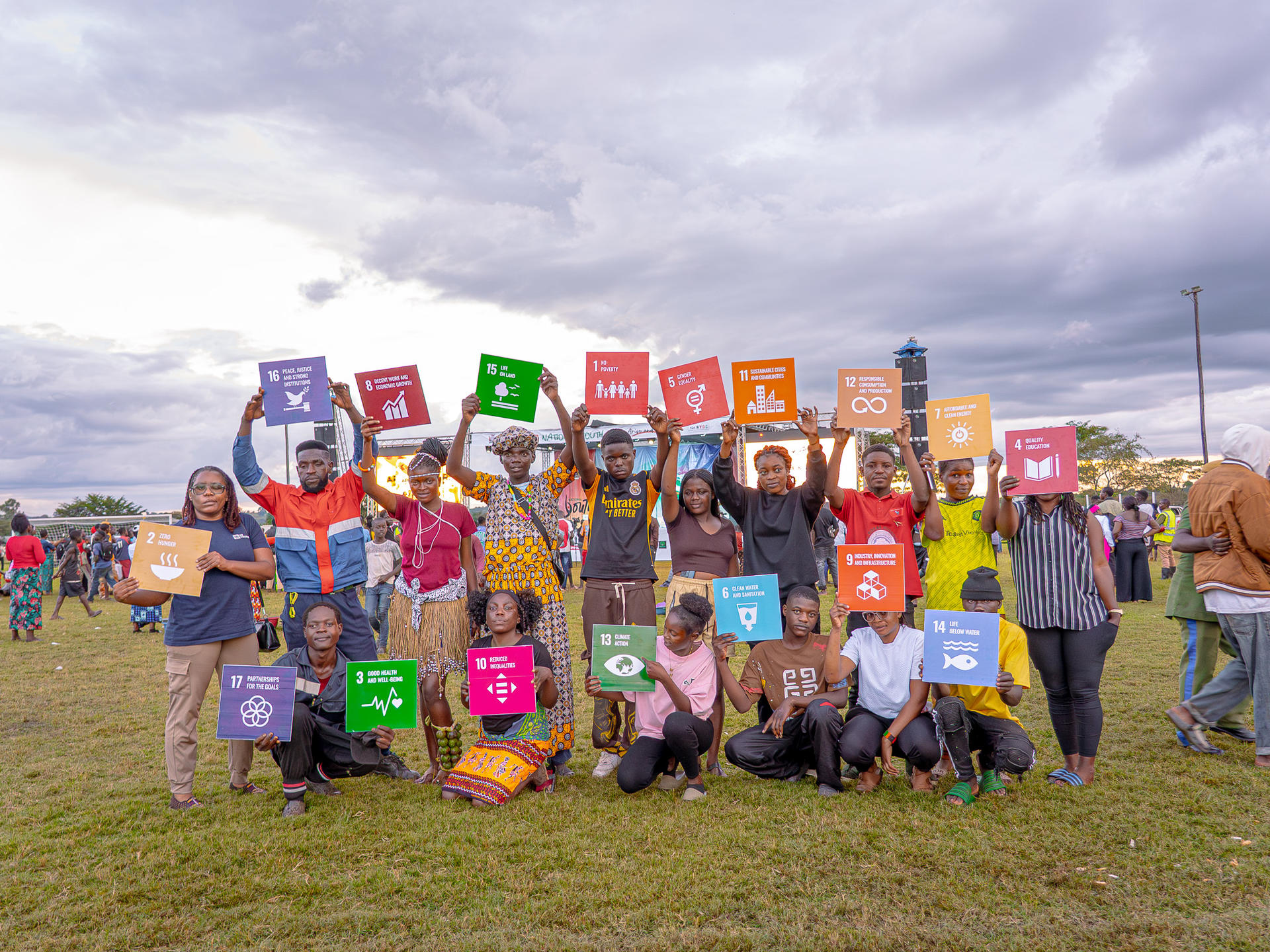 Group of people on a grassy field holding colorful square signs under a cloudy sky.