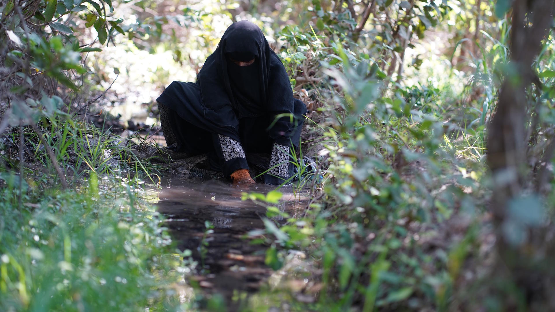 Person in a black full-body garment crouches beside a narrow water trough in a sunlit forest.