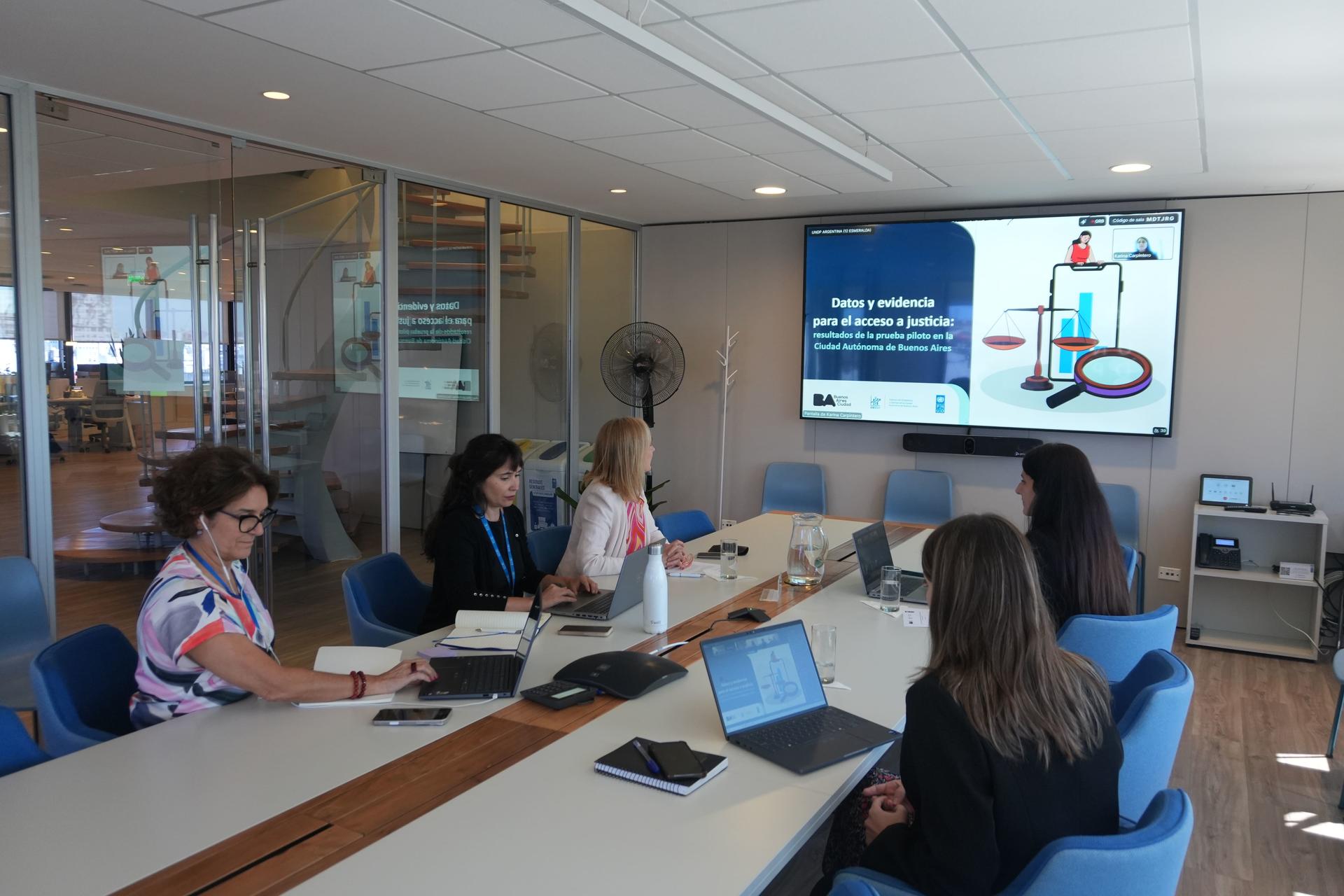 Masked team sits around a conference table in a modern office, slide on screen at front.