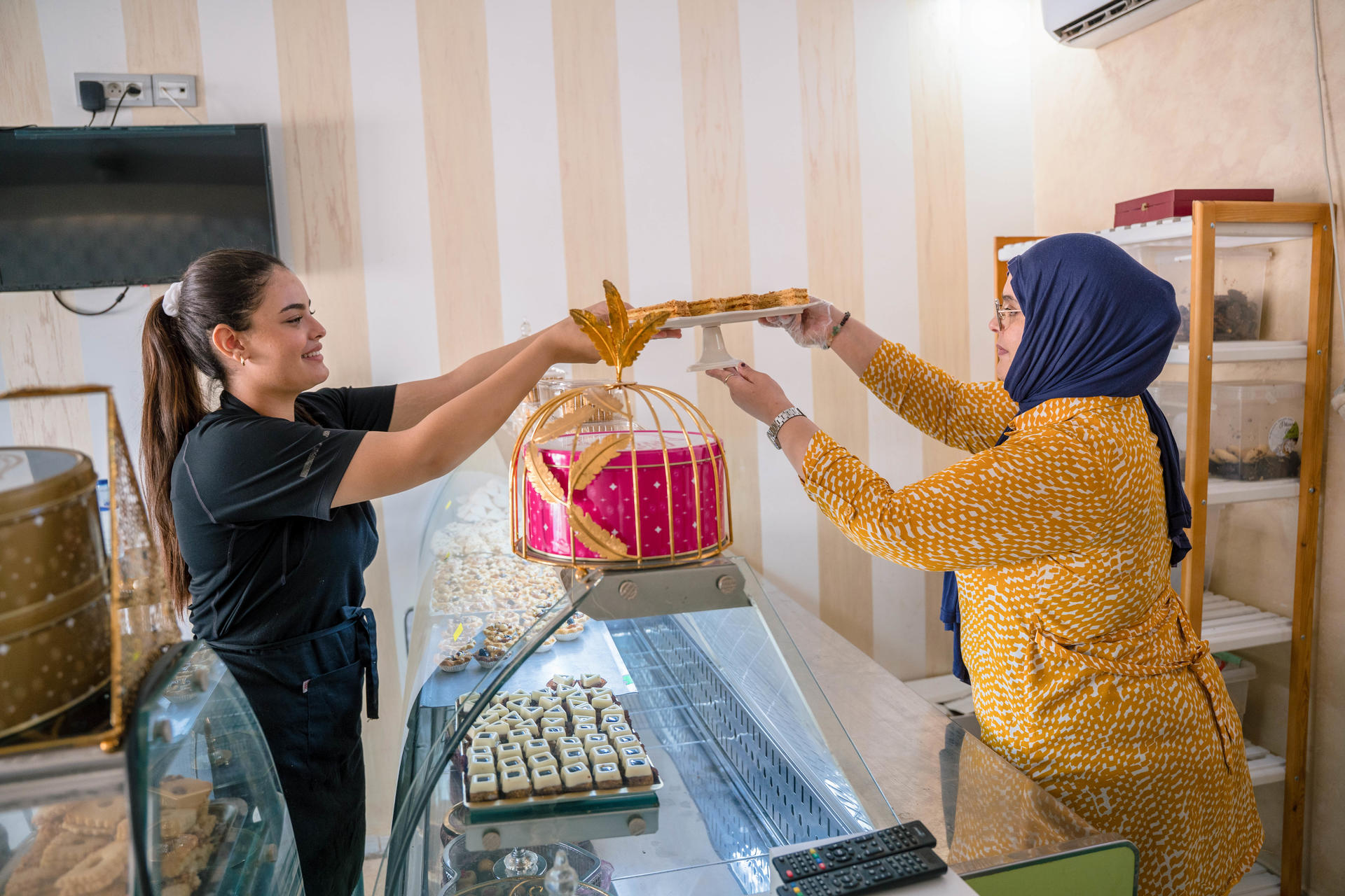 Two women in a bakery exchange a pink cake with a yellow ribbon over a glass pastry case.