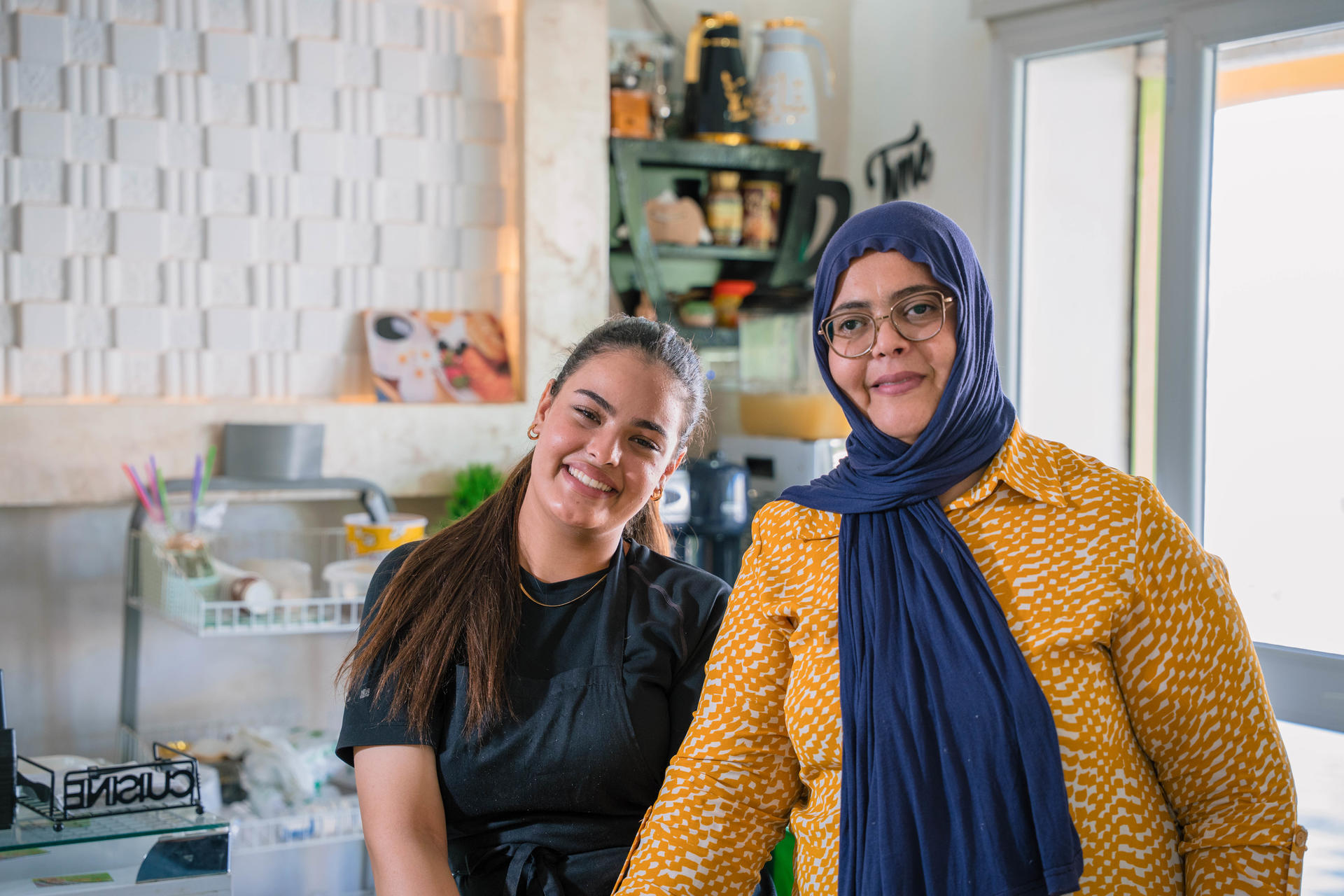 Two women in a bright kitchen; one in black, the other in a yellow jacket with a blue headscarf.