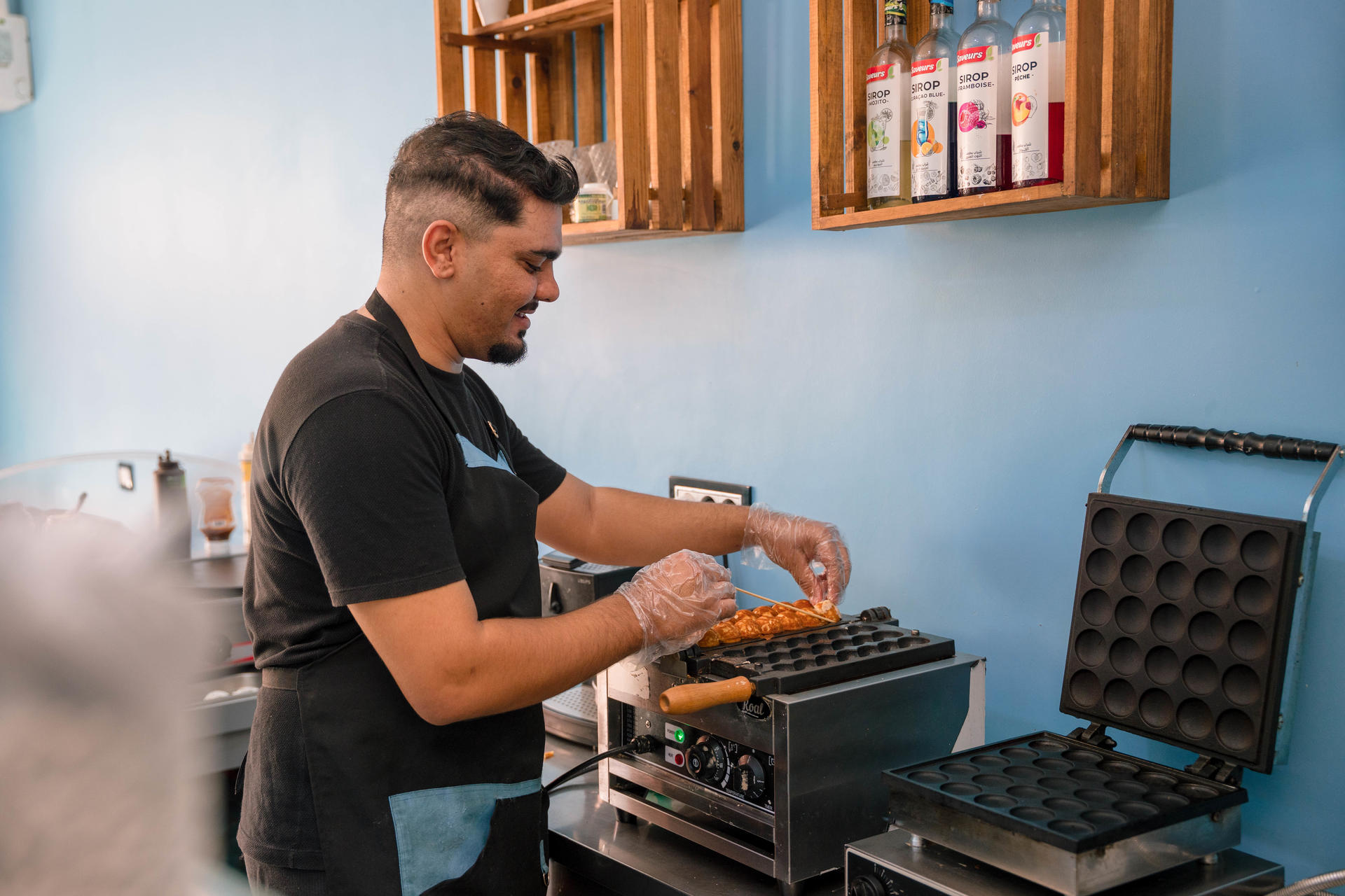 Person in a black shirt uses a waffle iron in a blue kitchen with bottle shelves.