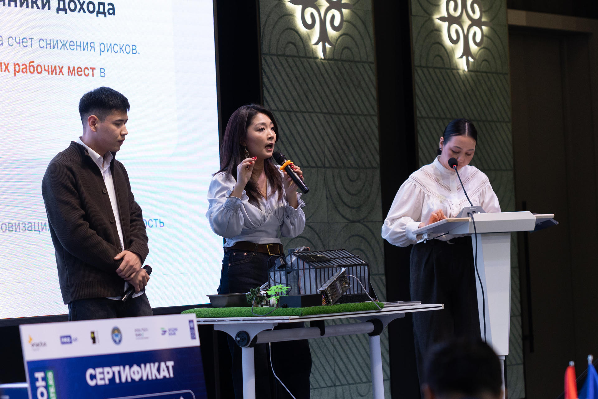 Three people on stage; a woman speaks into a microphone at center, flanked by two men, with a laptop and podium.
