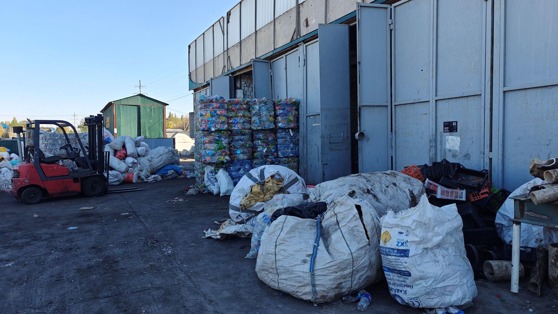 Industrial yard beside blue warehouse; red forklift, pallets, and large bags.