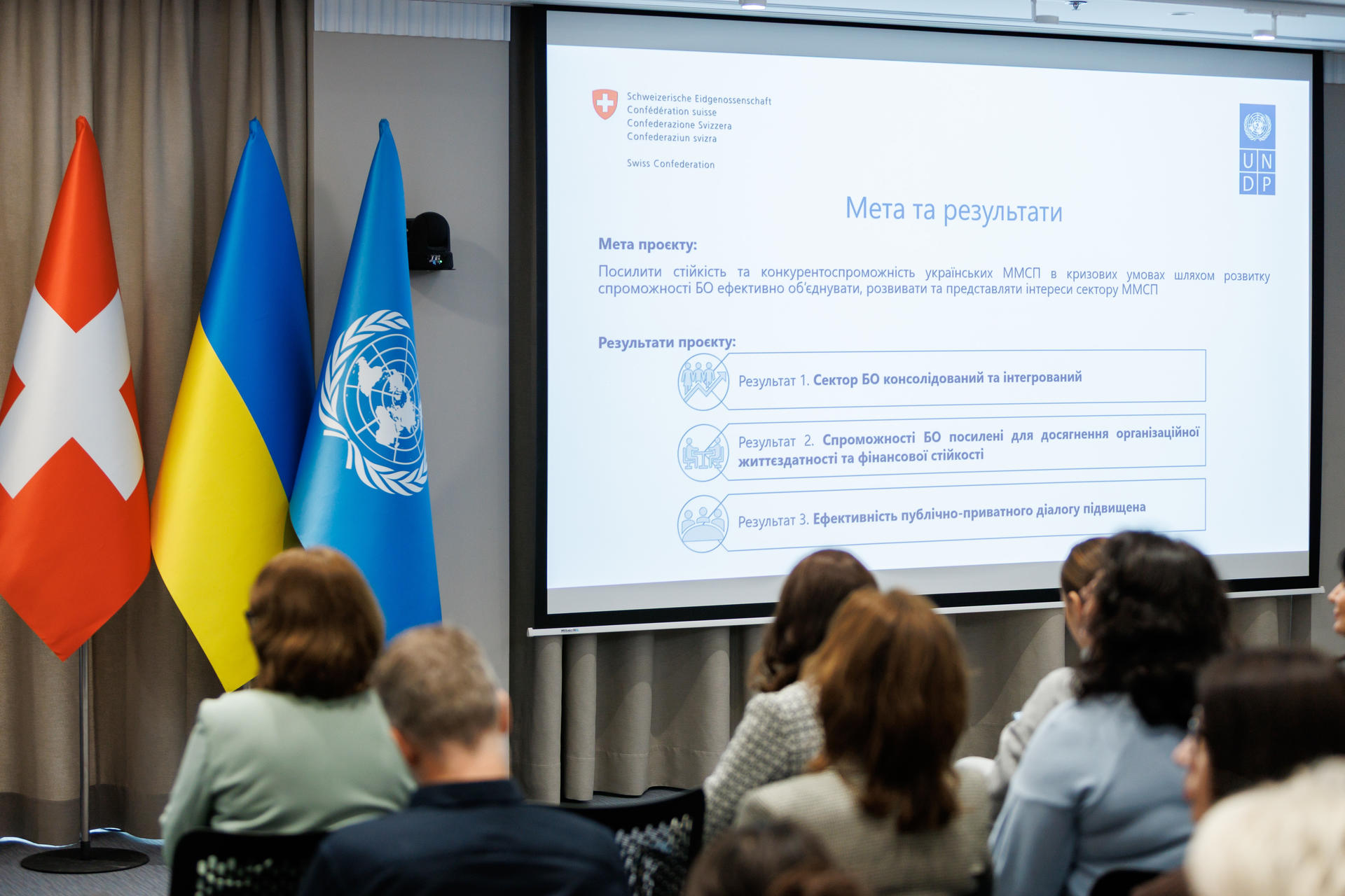 Audience in a conference room watches a slide with Swiss, Ukrainian, and UN flags beside screen.