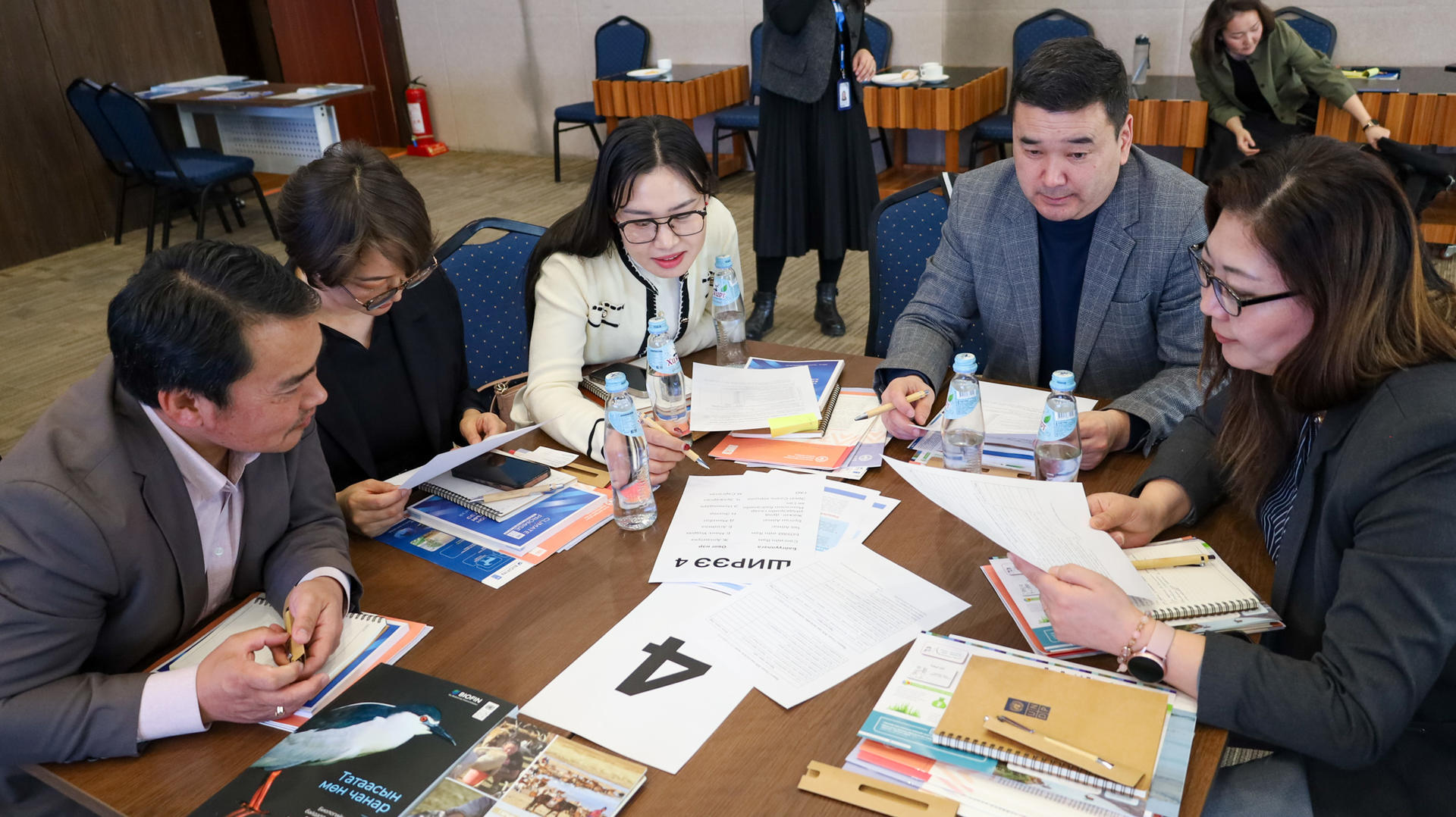 Six people sit around a table in a classroom, collaborating with papers, notebooks, and a laptop.