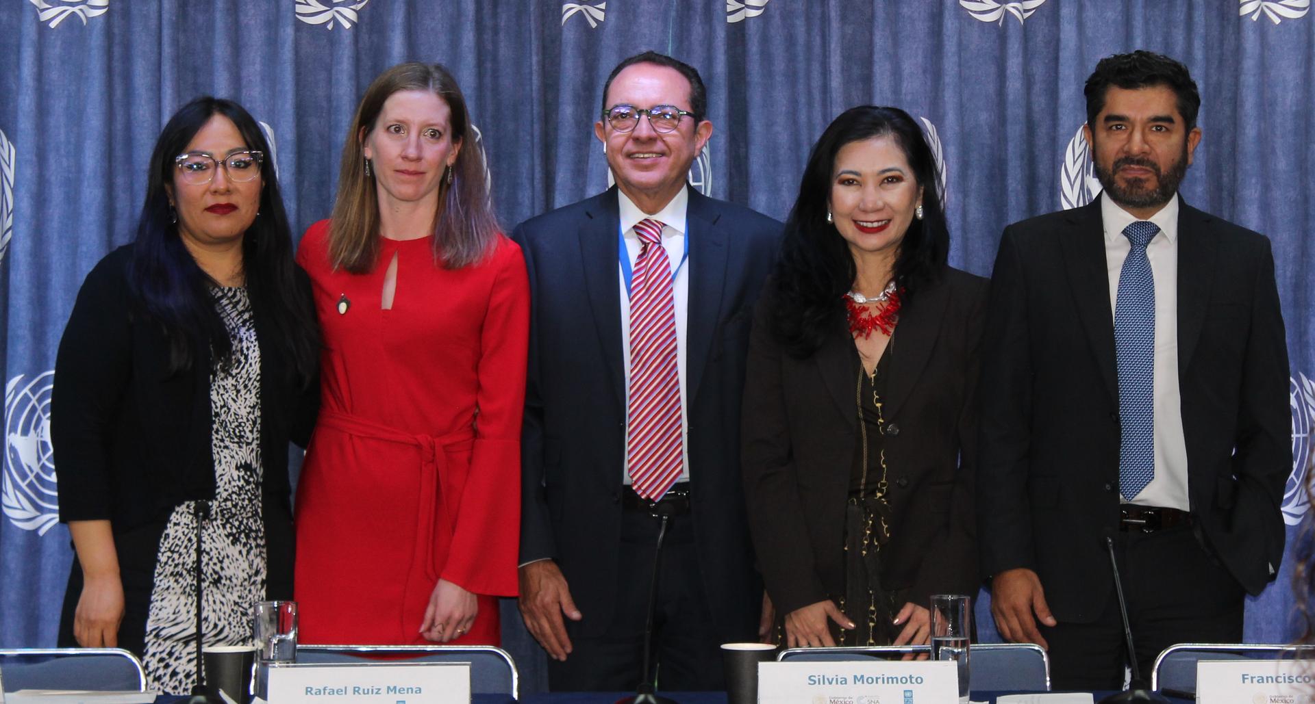 Six formally dressed people stand behind a table with nameplates before a blue curtain.