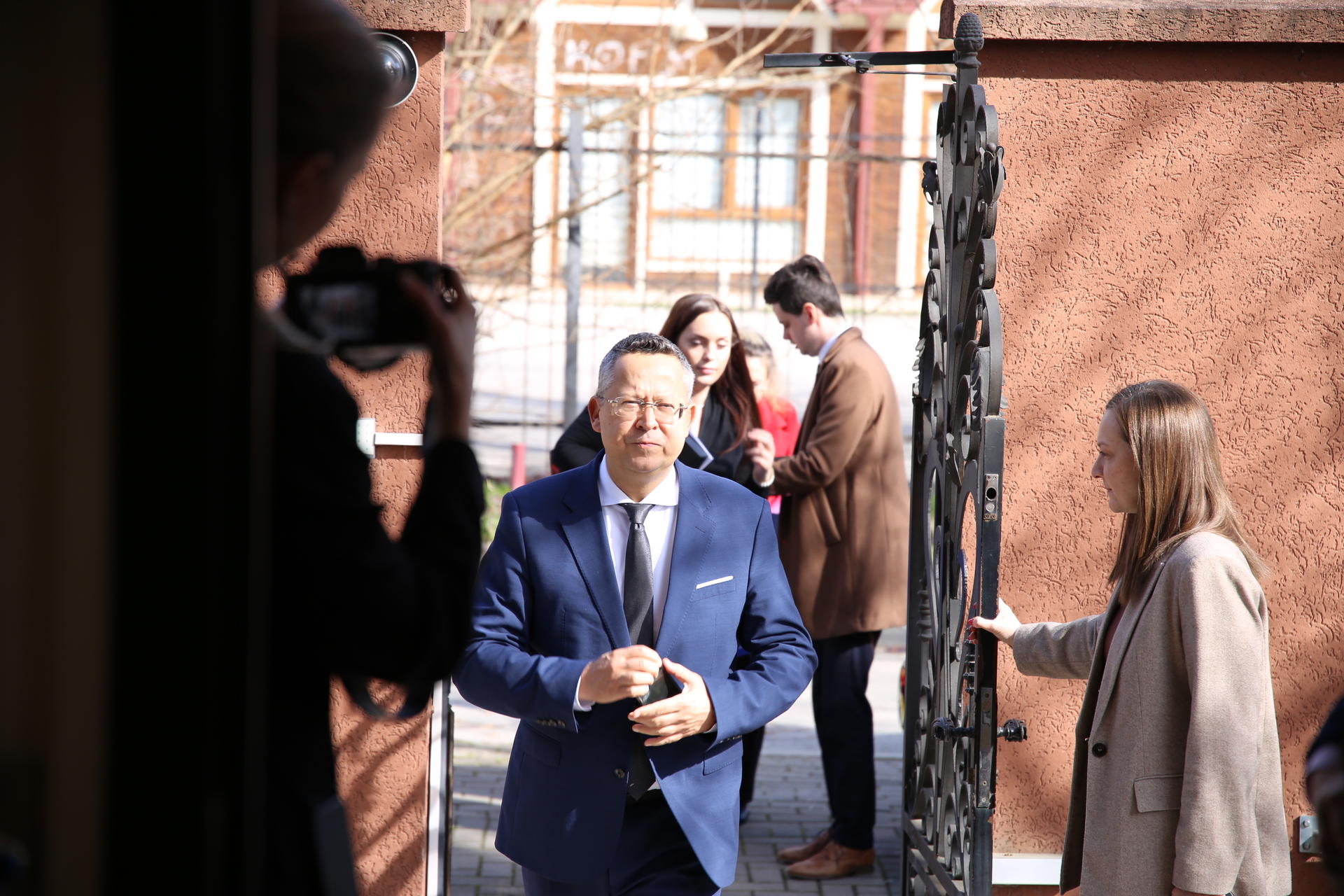 Man in a blue suit walks through a sunlit brick courtyard as photographers film him.