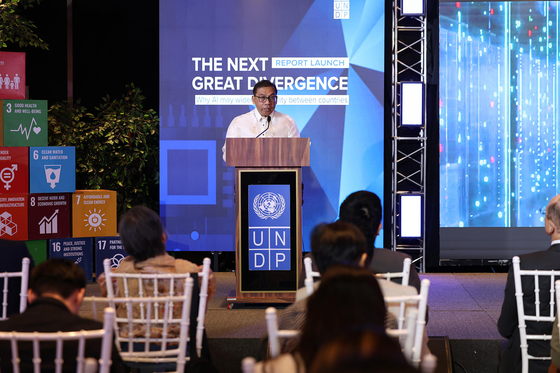 Speaker at a podium on a blue UNDP stage, audience seated; SDG blocks on the left.