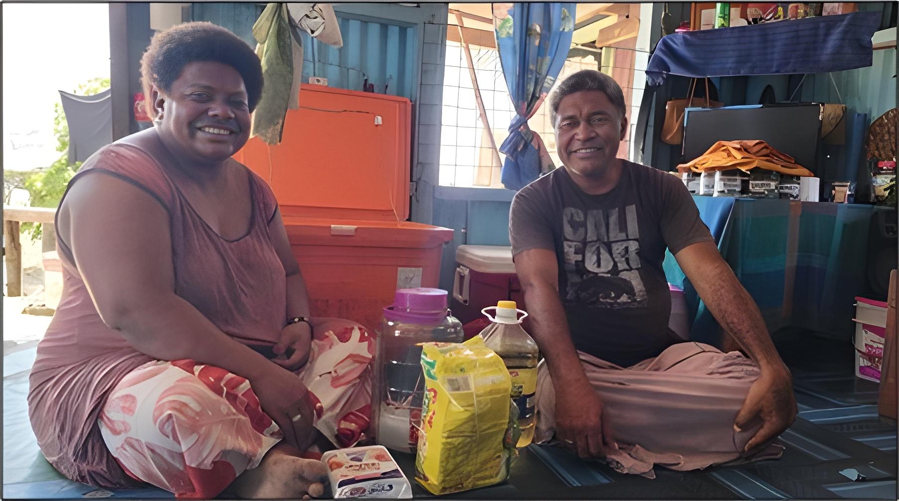 Two people sit on the floor in a shelter, surrounded by bags, a yellow bottle, and orange crates.