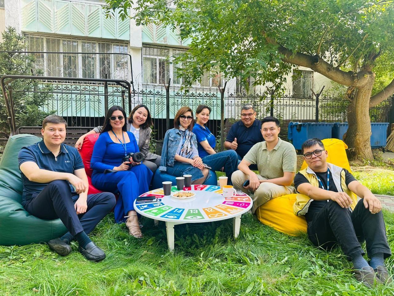 Group photo: people sitting on grass around a colorful round table outdoors, smiling.