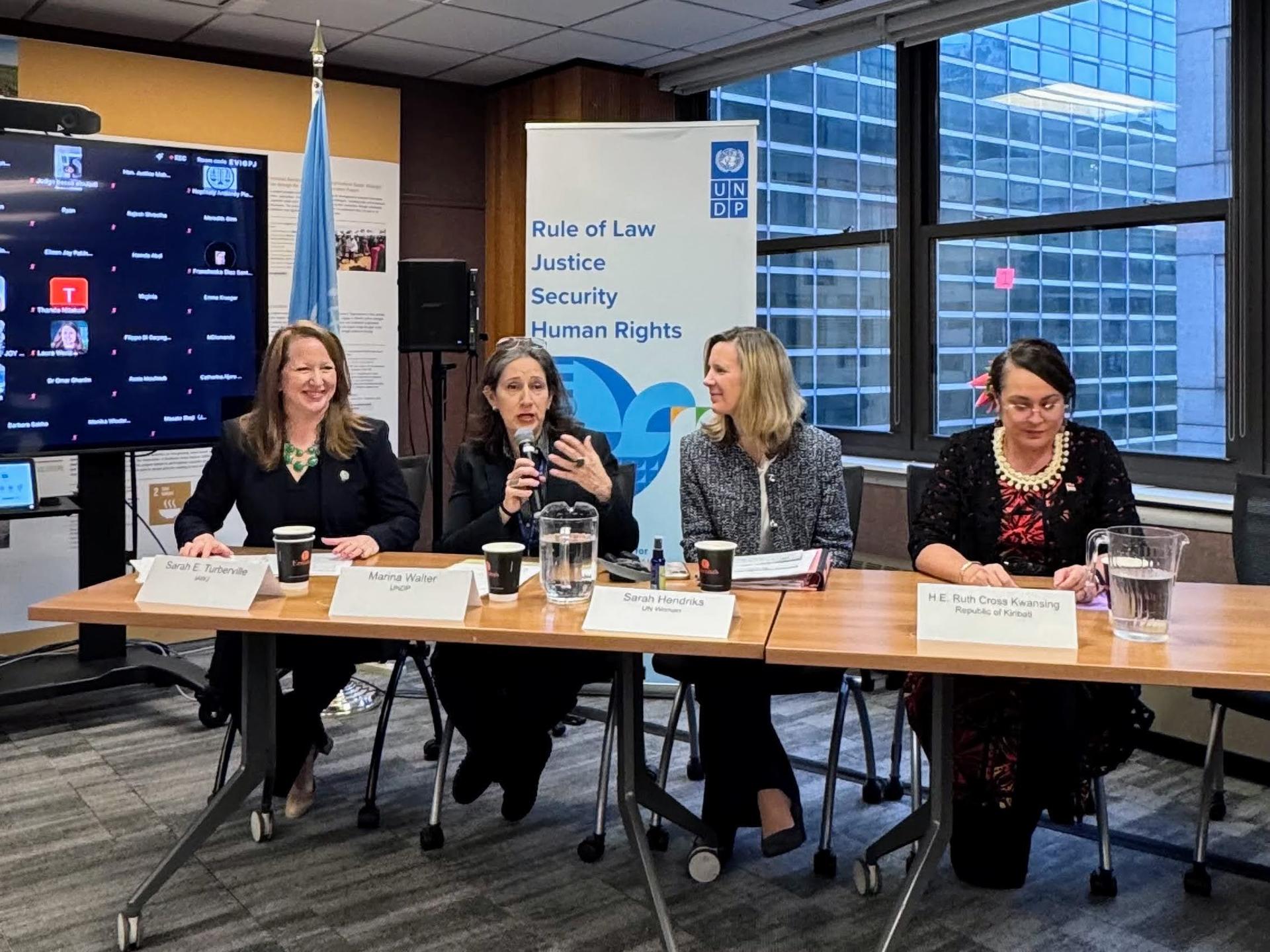 Four women panelists sit at a long table with microphones and water, conference banner behind.