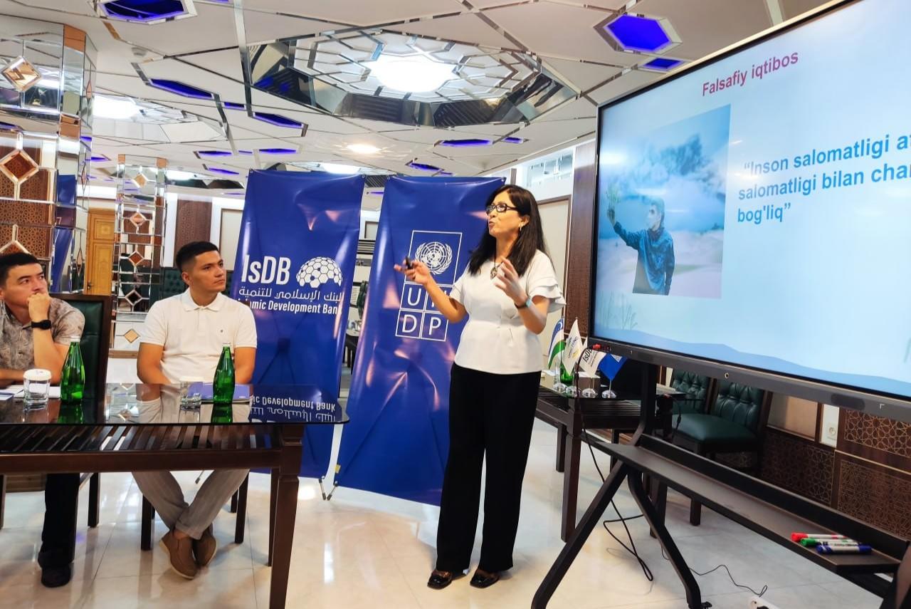 Photograph of a woman in a white top presenting at a booth with blue banners and a large screen.