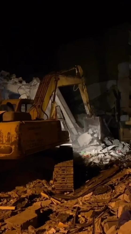 Nighttime construction site with an orange excavator scooping rubble under artificial lights.