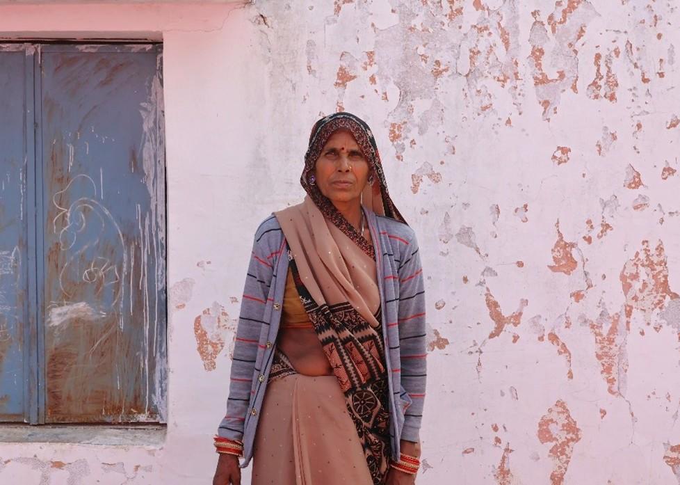 Person in a brown hijab and tan coat, back turned, beside a weathered white wall and blue door.