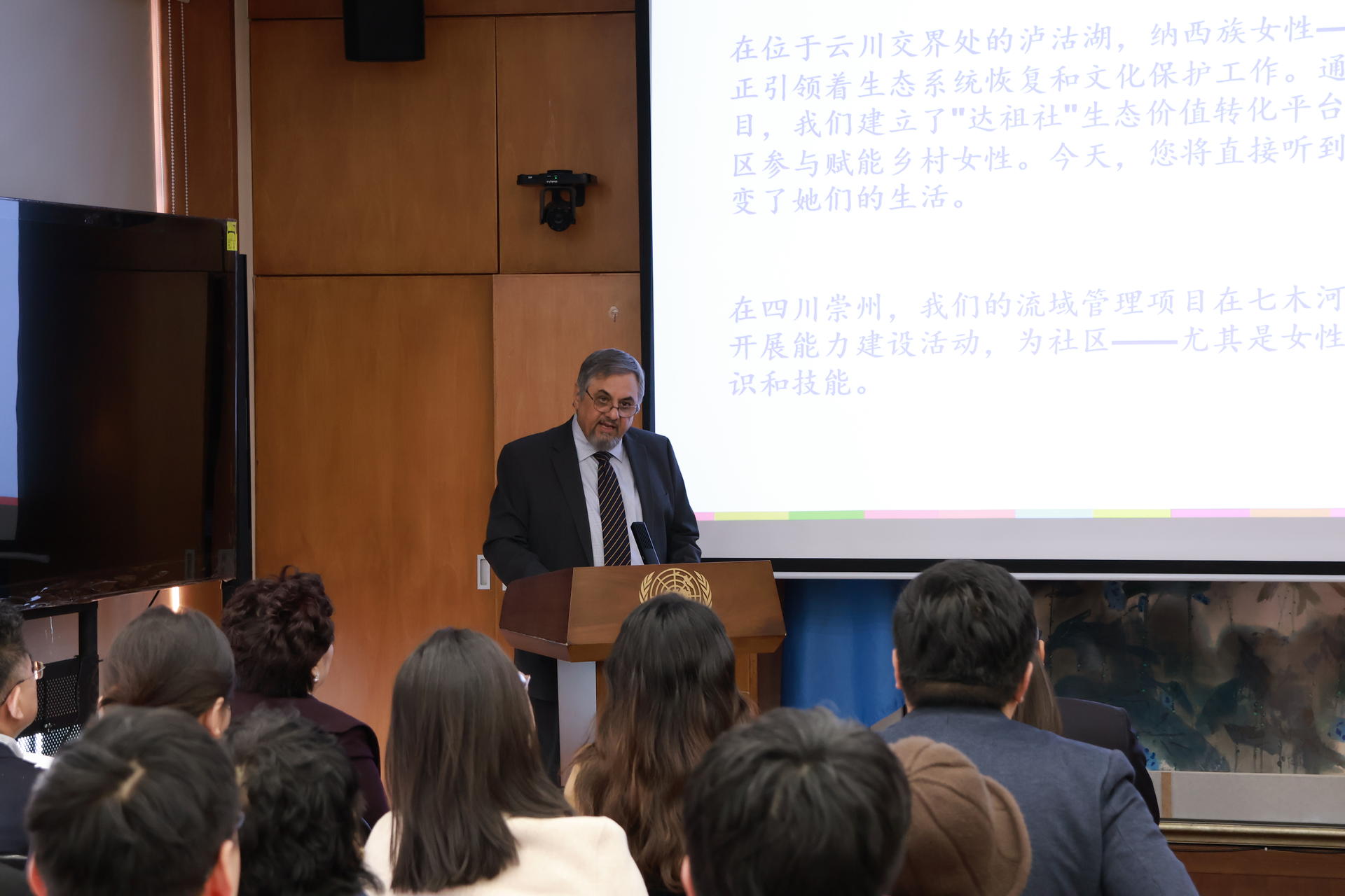 Man in a suit presents at a podium to an audience, with a projected slide behind.