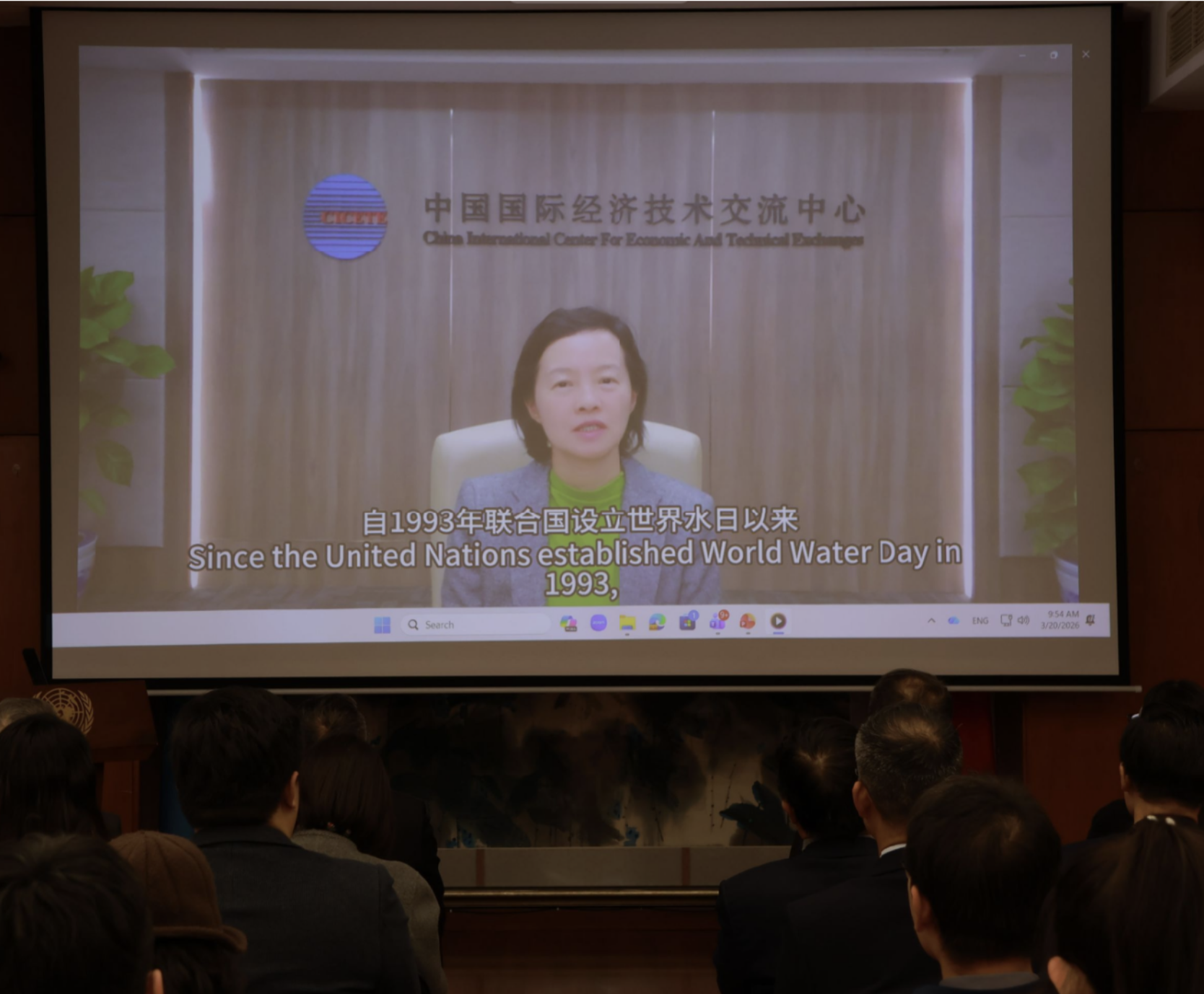 Audience seated in a conference room watching a large screen showing a presenter and subtitles.