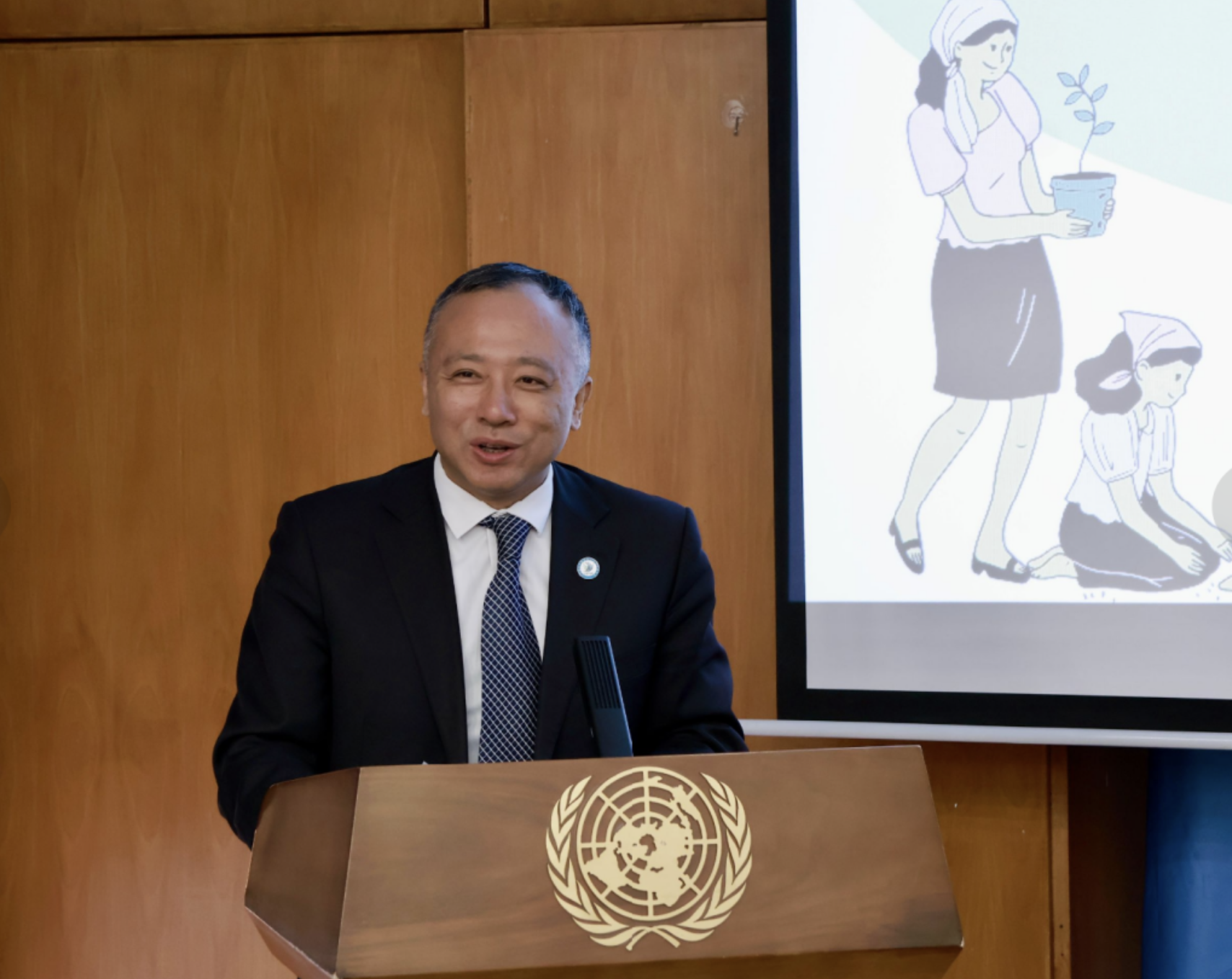 Man in a dark suit at a United Nations podium in a wood-paneled room; cartoon projected.