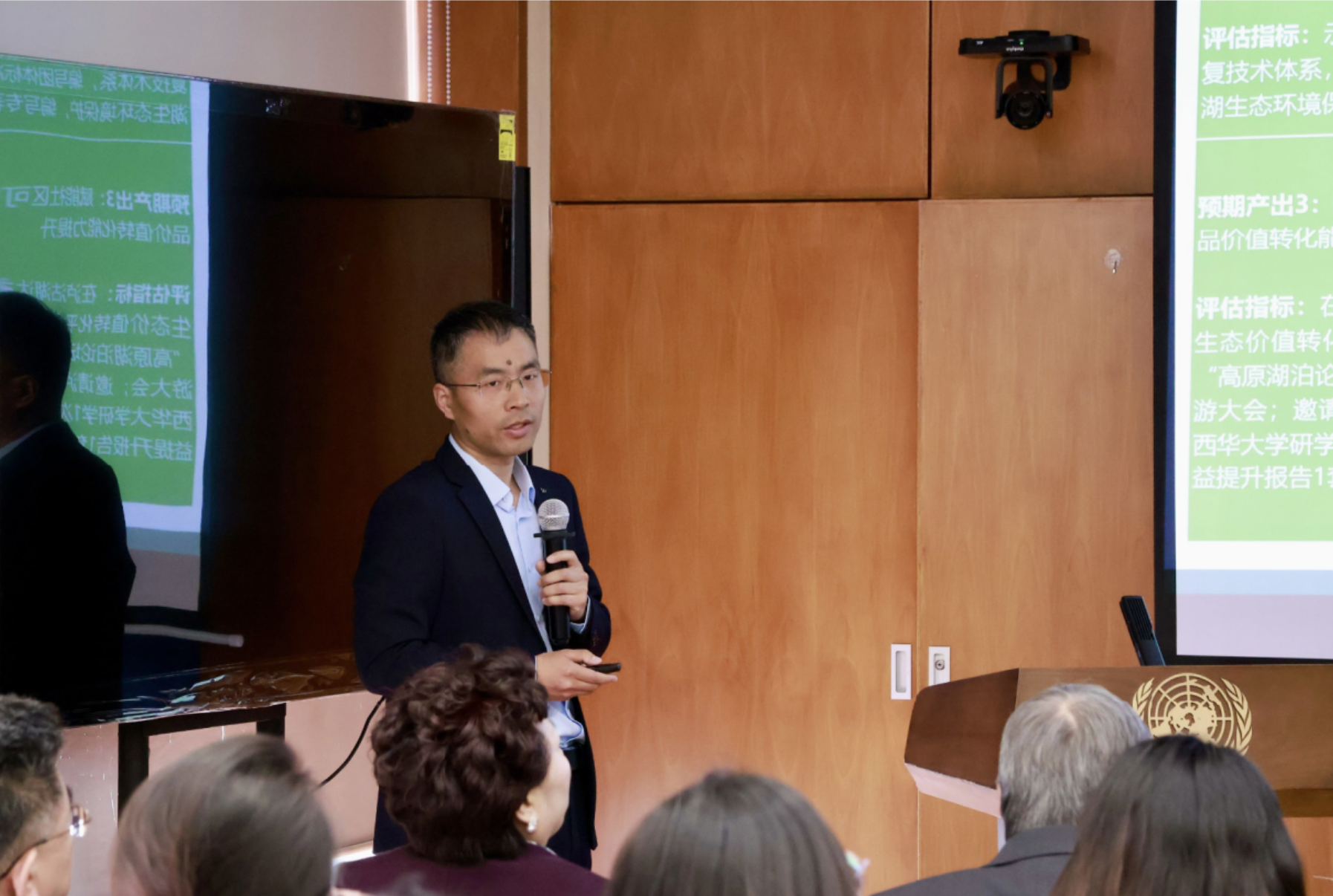 Photograph: man in a suit speaks into a microphone in a lecture room, green slides on projector.